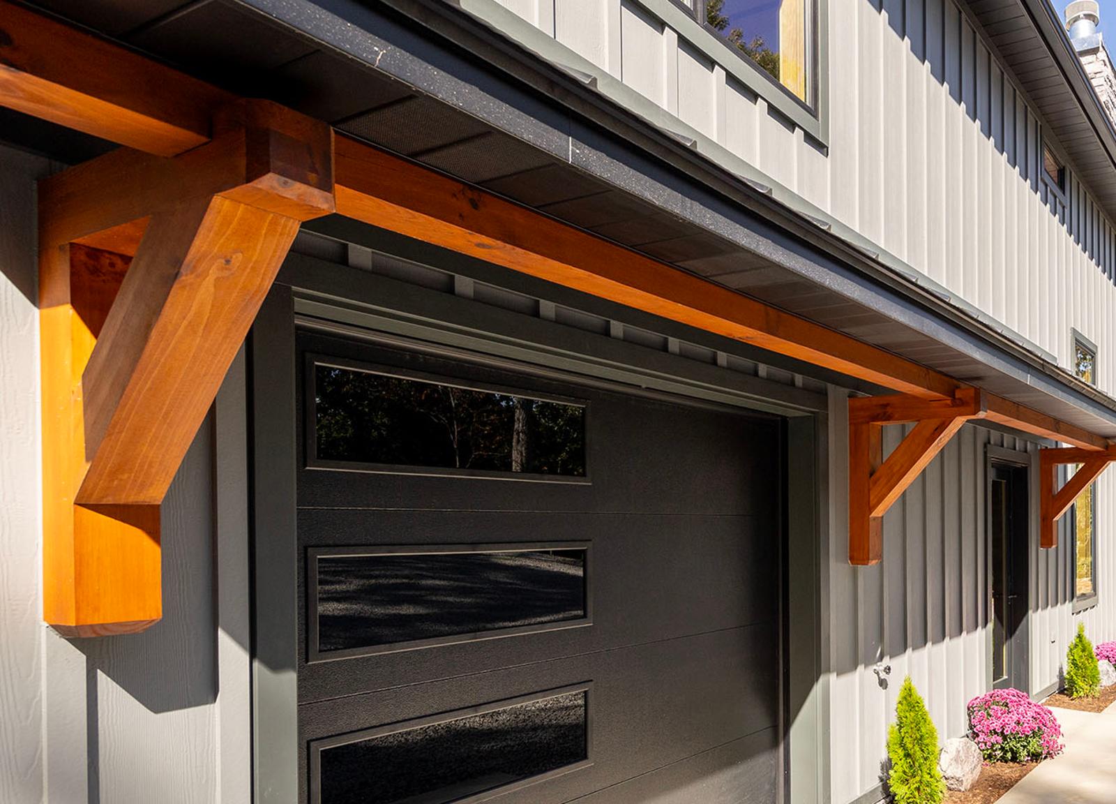 Close view of a stained timber knee brace bracketed under a roof eave against dark board-and-batten siding, above a black garage door
