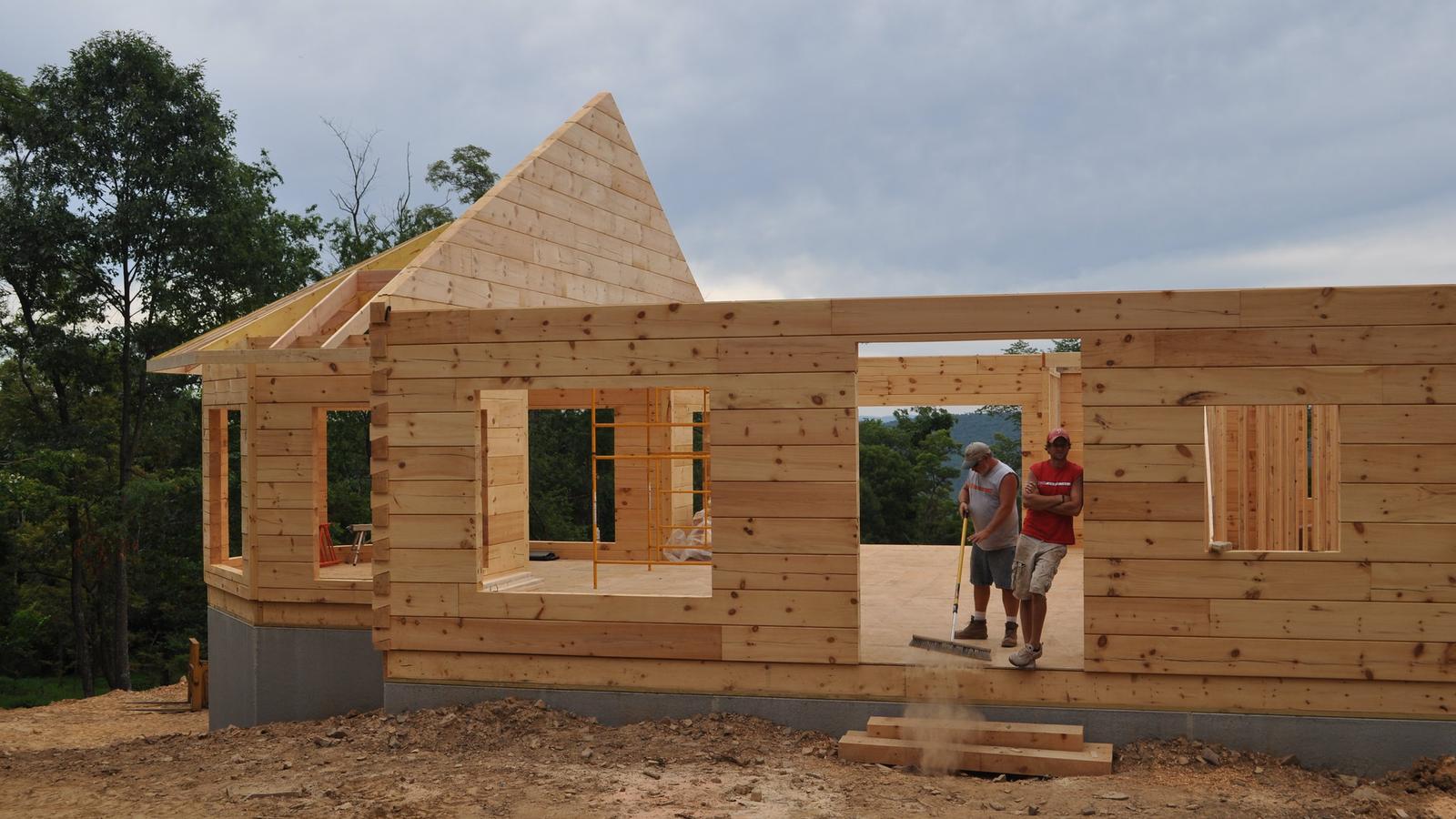 A half-built log house on a graded ridgetop site: stacked log courses, four open window frames, a gable truss sheathed and standing, and two of the crew at work through a door opening
