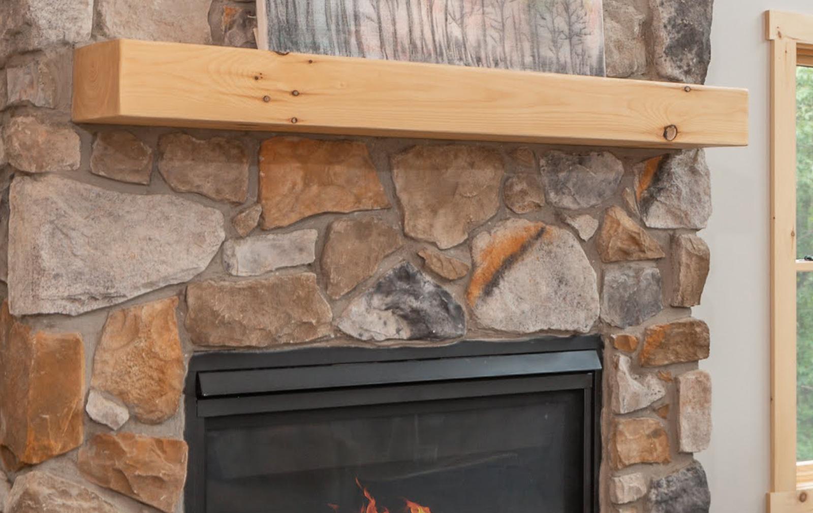 Close view of a squared timber mantel with visible knots spanning a chimney breast of irregular tan and gray fieldstone set with wide mortar joints, the top of a lit firebox below