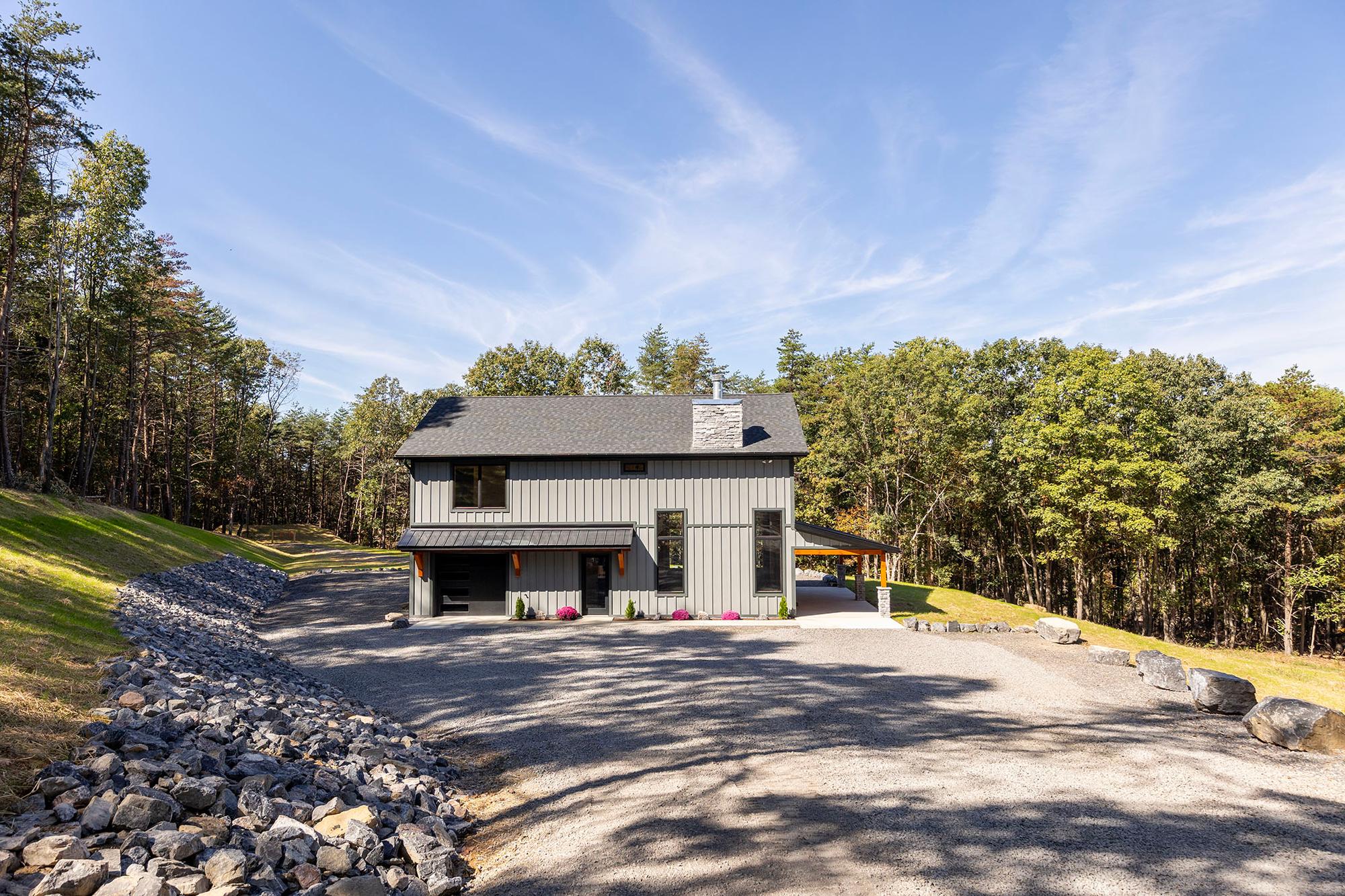 Two-story home with gray board-and-batten siding, stone chimney and covered patio, fronted by a wide gravel drive edged with rock