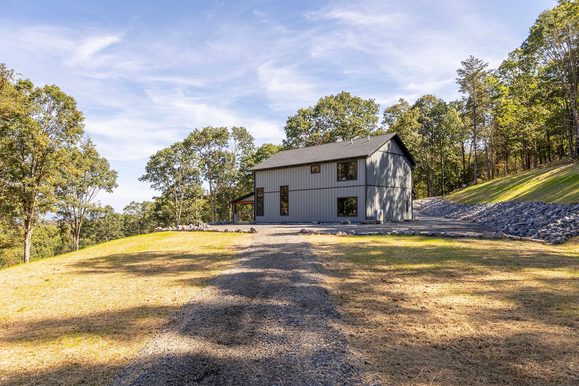 The home on its hilltop clearing seen from below, a gravel path and rock-lined bank rising toward the covered patio