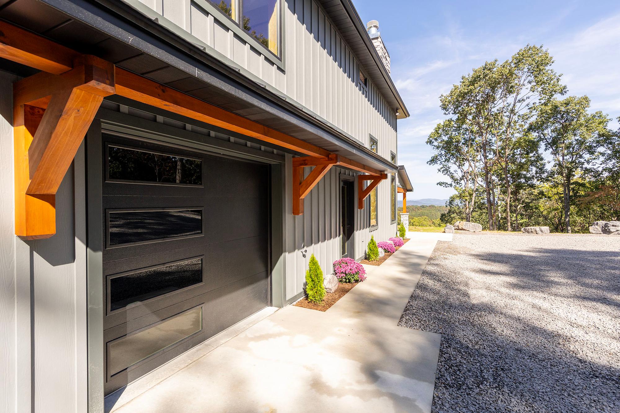 Black glass-paneled garage door under timber knee braces, with mums and young evergreens planted along the concrete walk