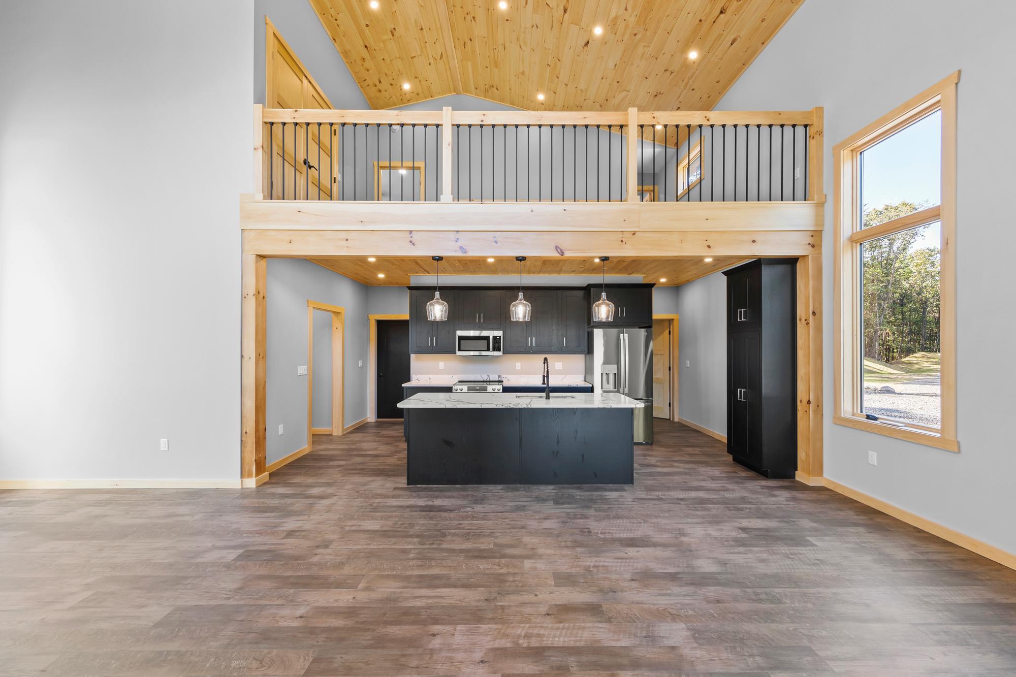 Empty great room looking toward the kitchen, with a vaulted pine ceiling, iron-spindle loft rail and a marble-look island