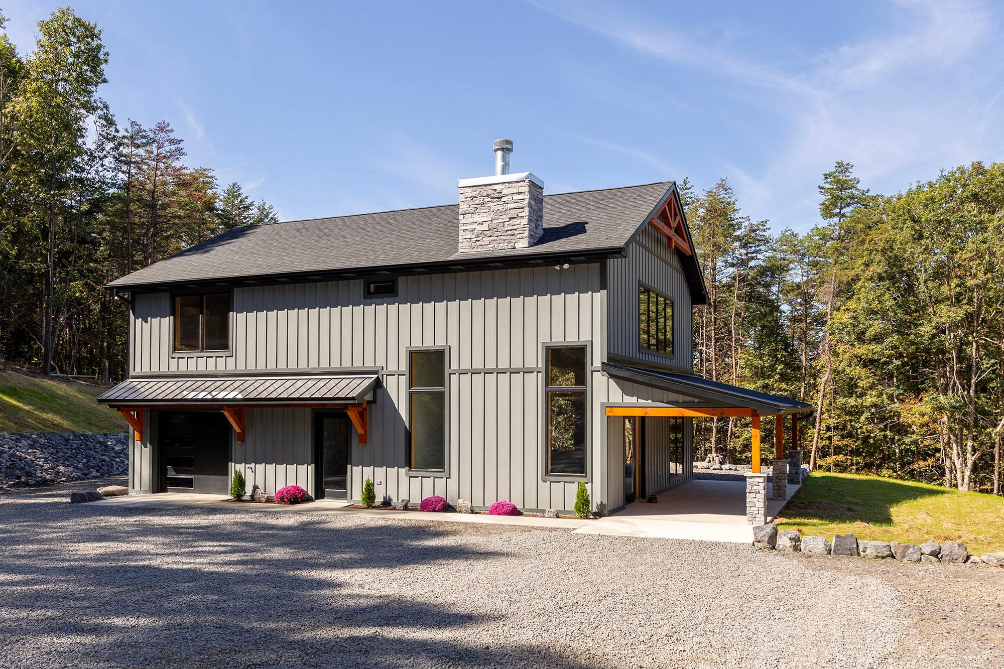 Gray board-and-batten home with a stacked-stone chimney, timber gable truss, covered entry, and flowering mums along the concrete walk