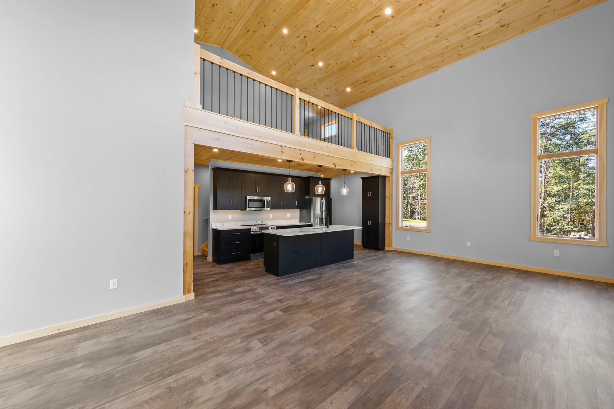 Unfurnished great room with tall windows into the woods, the kitchen and loft beyond, and gray plank flooring throughout