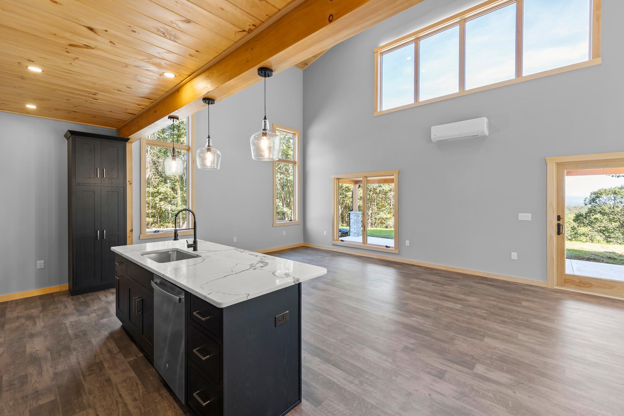Kitchen island with sink and stainless dishwasher facing the empty great room, a mini-split high on the wall and glass door beyond