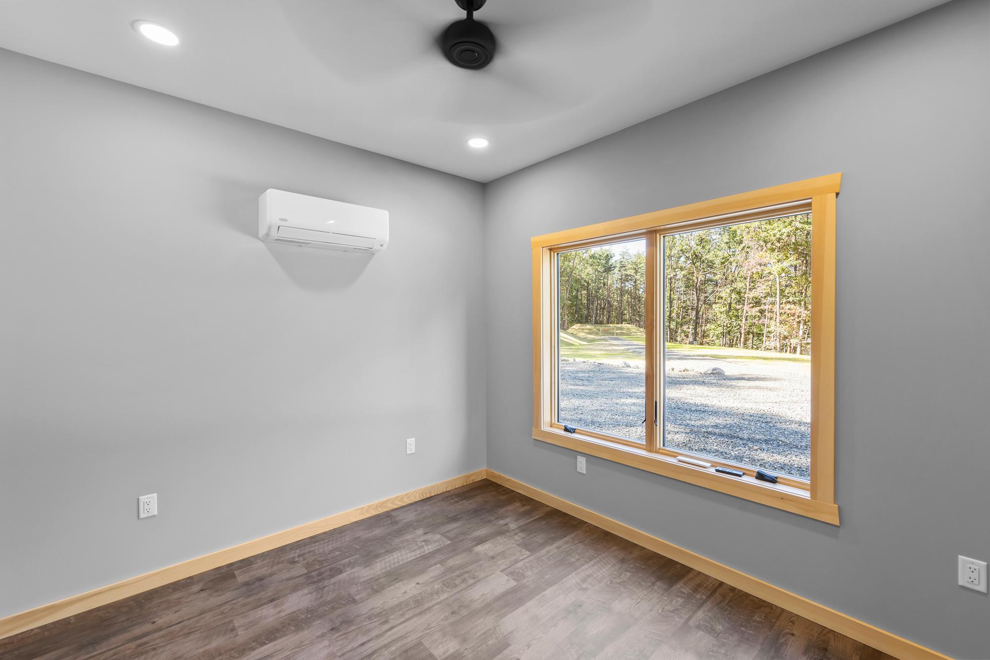 Empty bedroom with a ceiling fan, wall-mounted mini-split and a pine-trimmed window onto the gravel drive and woods
