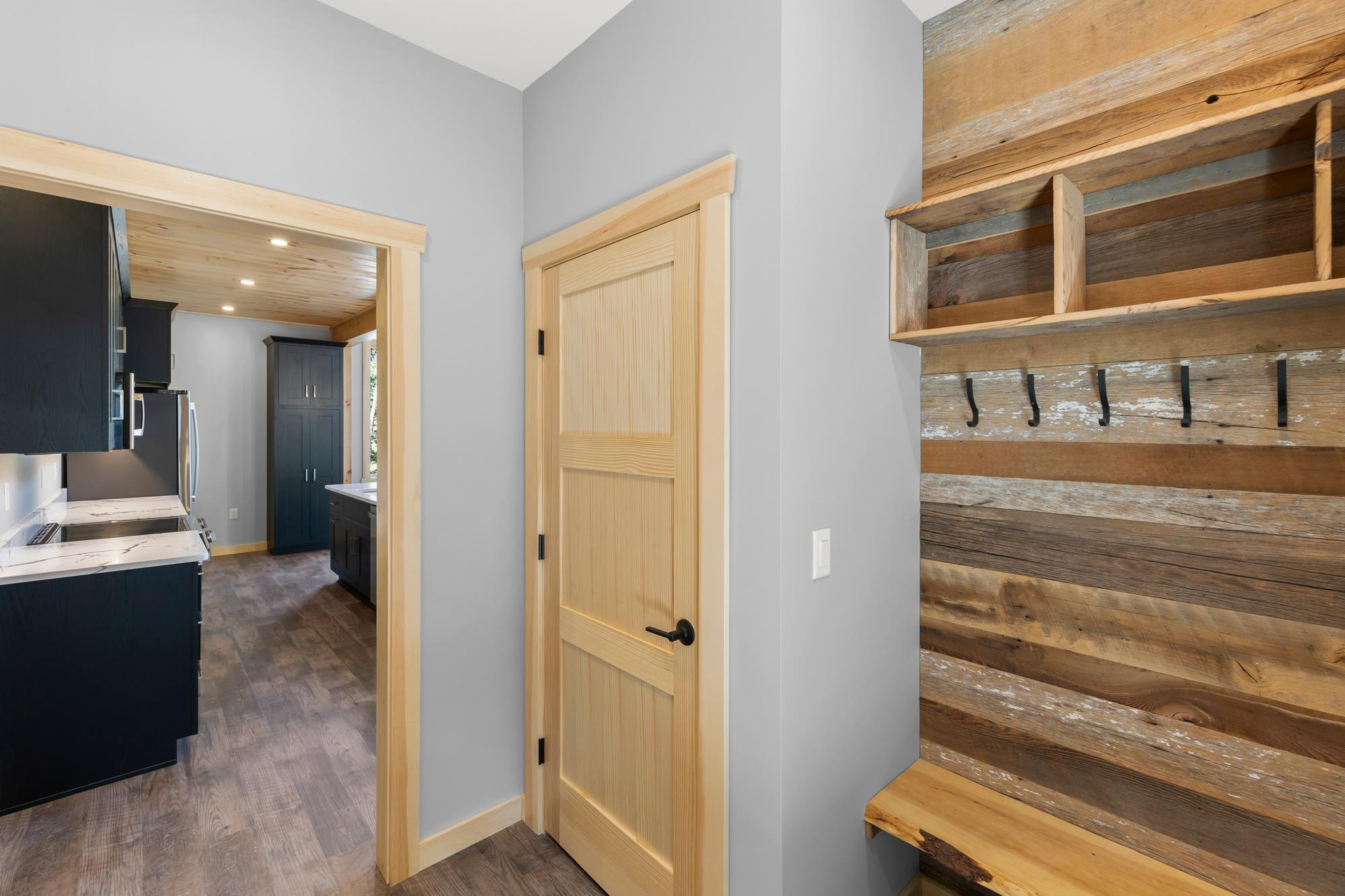Mudroom wall of reclaimed barnwood with black coat hooks, a cubby shelf and live-edge bench, the kitchen visible through the doorway