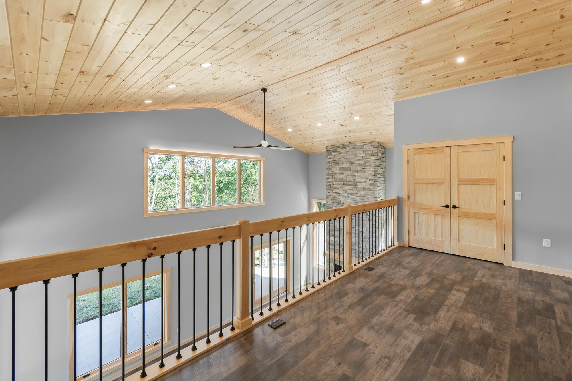 Loft with a plank pine ceiling, timber-and-iron railing, the stacked-stone chimney rising past it, and double pine doors to a bedroom