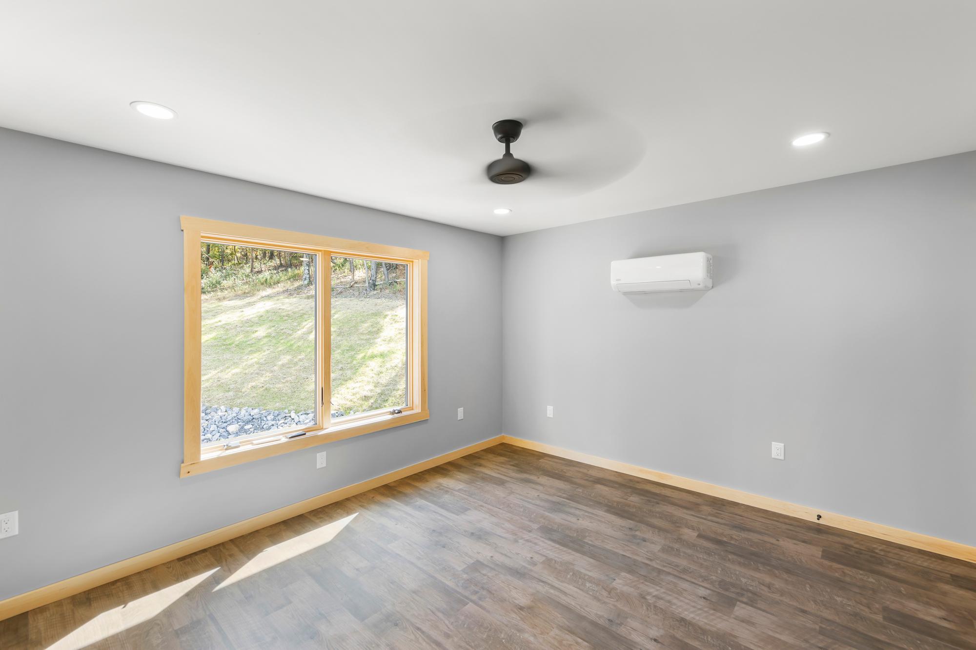 Bare bedroom with a window onto the grassy slope and rock bank, a mini-split on the wall and ceiling fan overhead