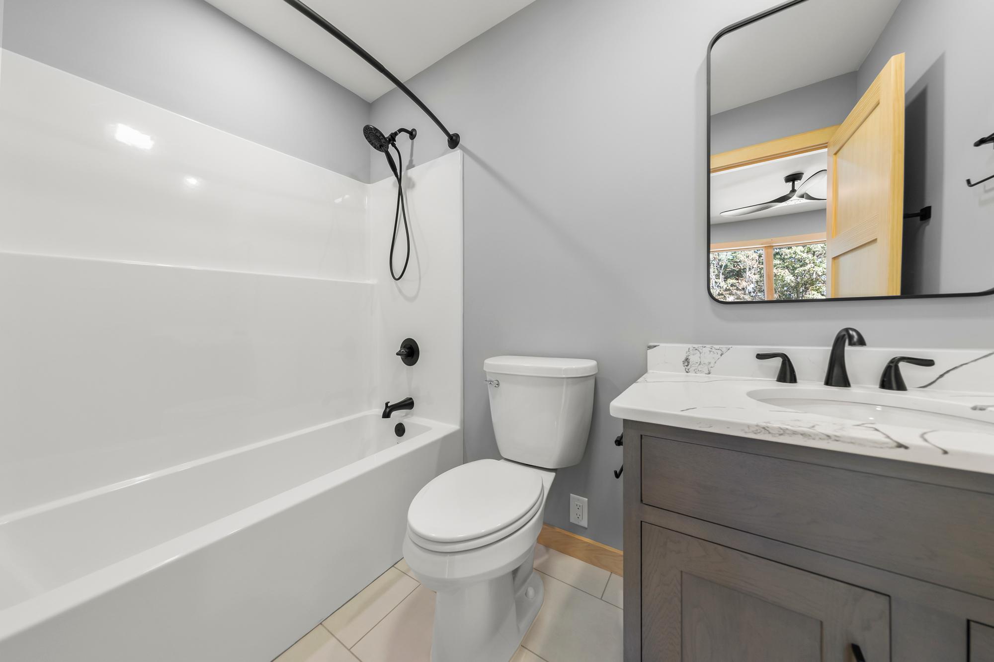 Hall bathroom with a white tub-shower, black hand shower on a rail, toilet, and dark vanity under a rounded black mirror