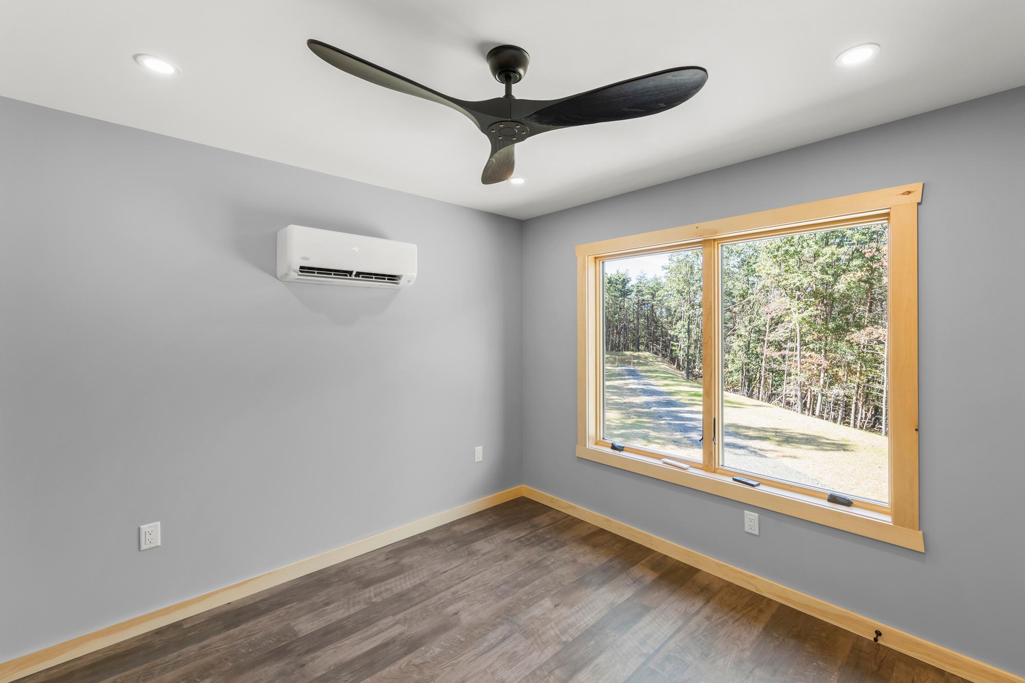 Empty bedroom with a black three-blade ceiling fan, a mini-split, and a window looking down the gravel drive through the trees