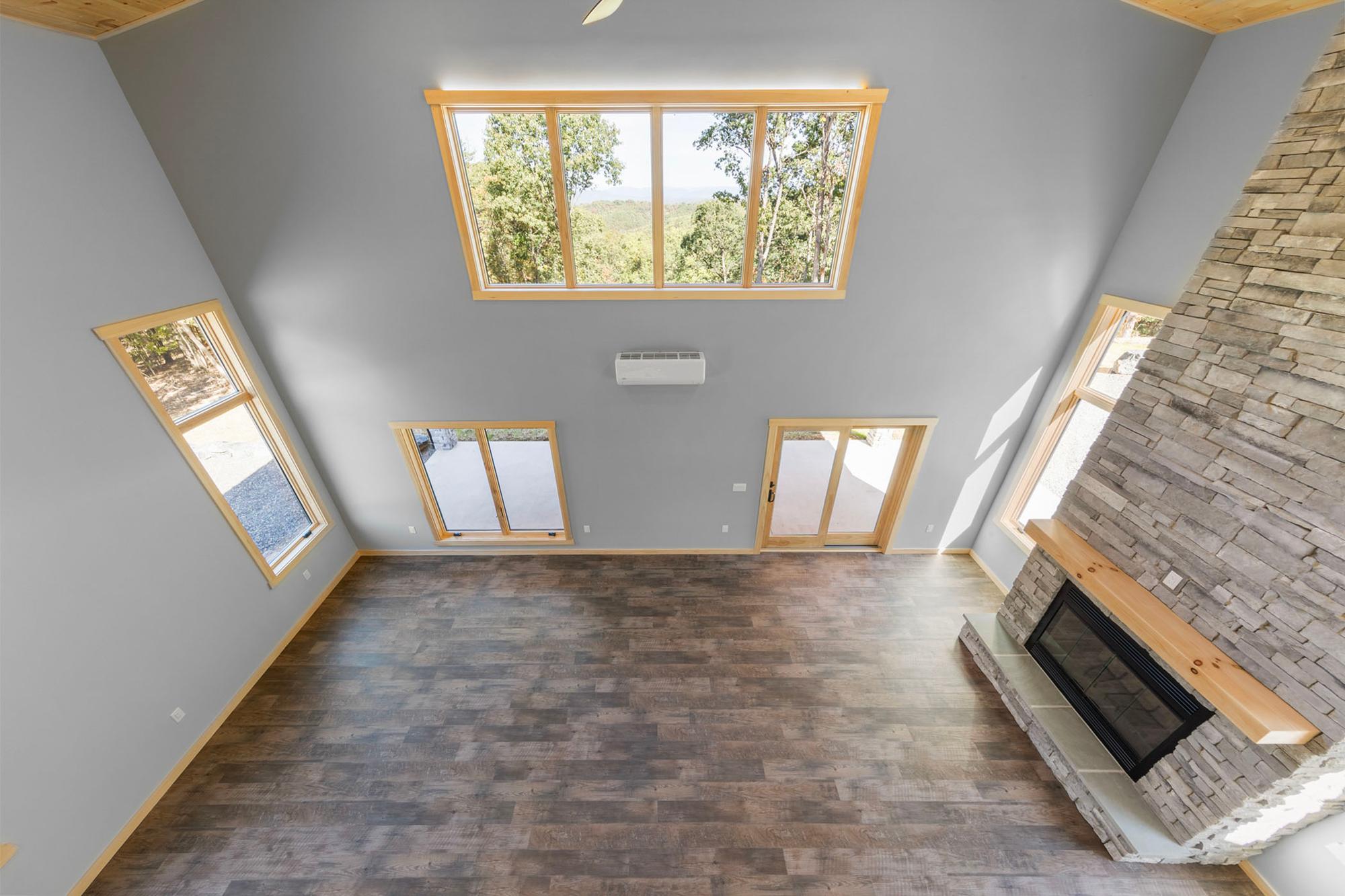 Great room seen from the loft — bare floor, a window band with wooded-hills view, mini-split, glass doors and the stone fireplace