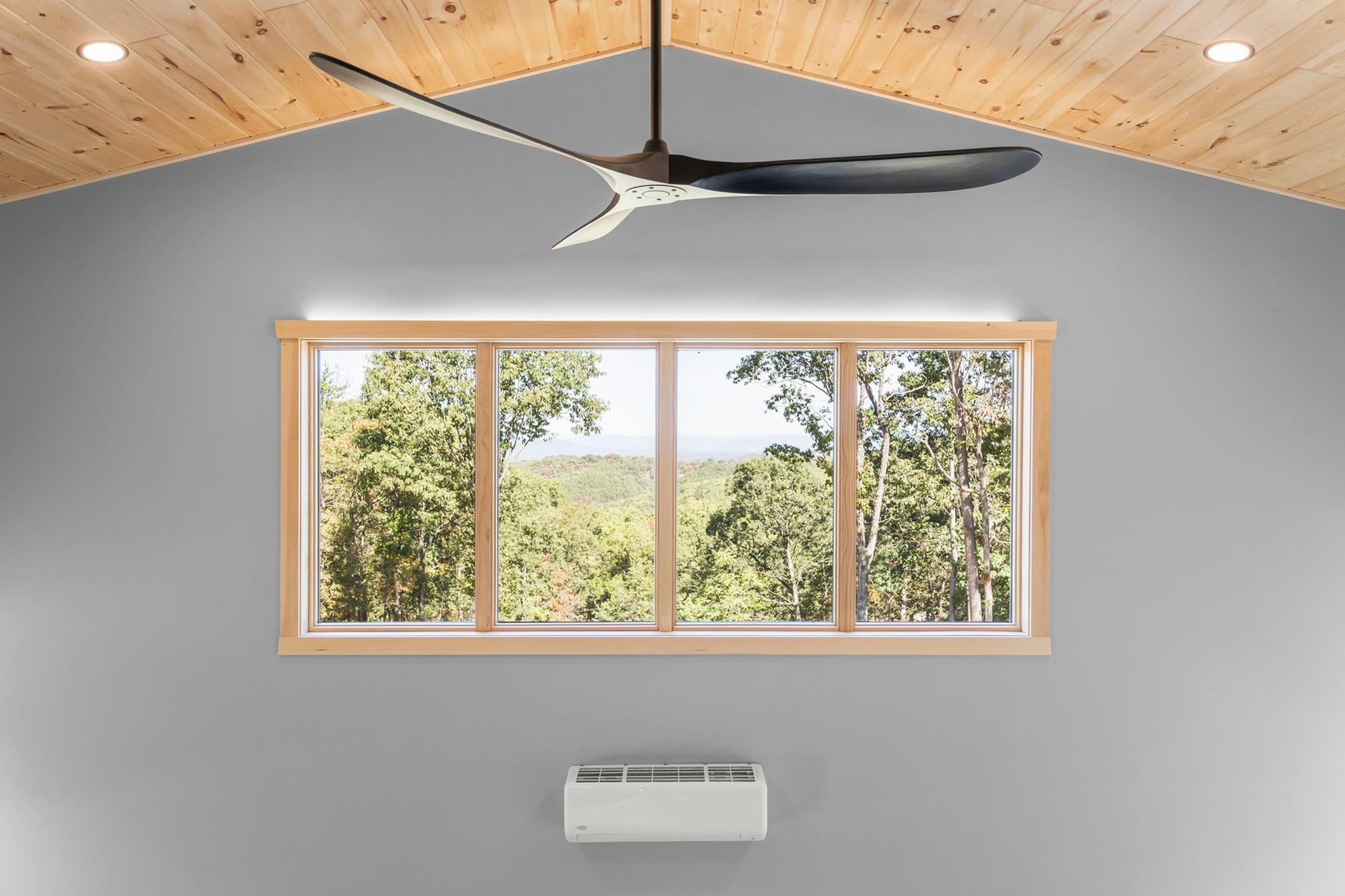 Four-pane window framing treetops and distant ridges under the peak of the pine ceiling, a mini-split centered below