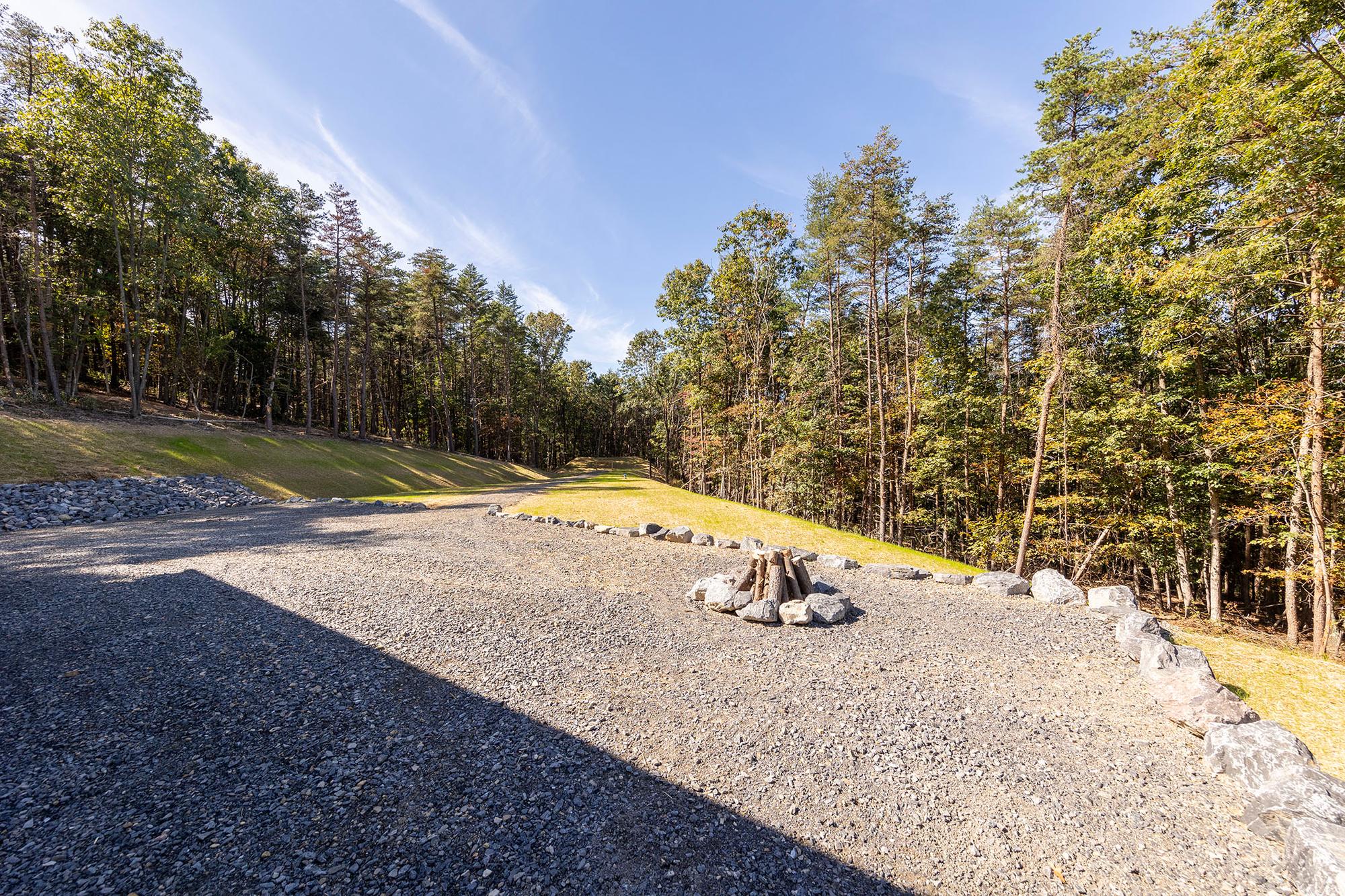 Gravel turnaround with a boulder-ringed fire pit stacked with logs, beside a mowed strip running between two tree lines