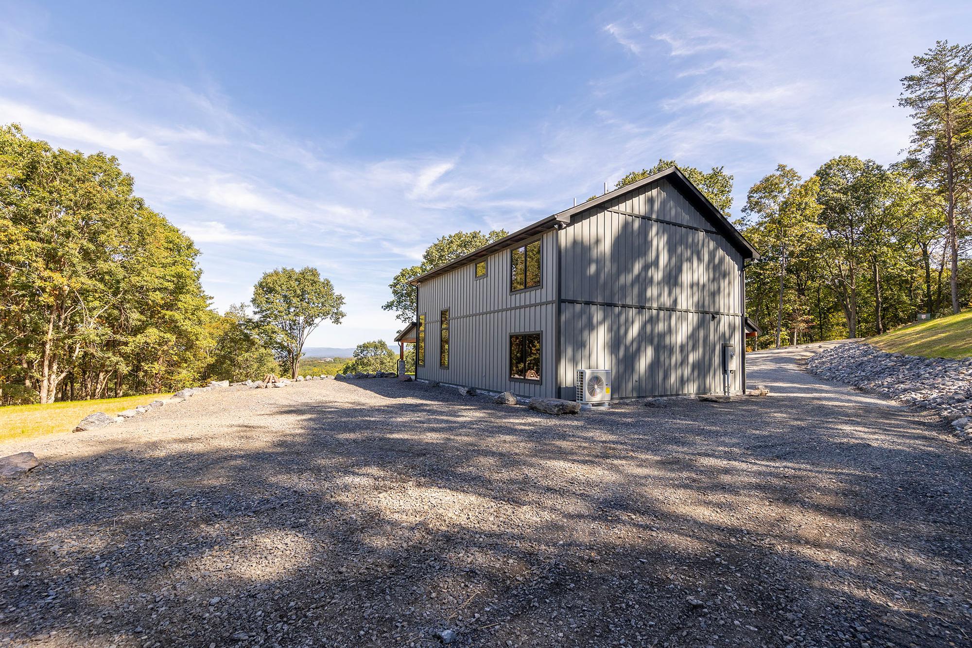 The home seen across the wide gravel parking area, a mini-split condenser against the wall and woods on three sides