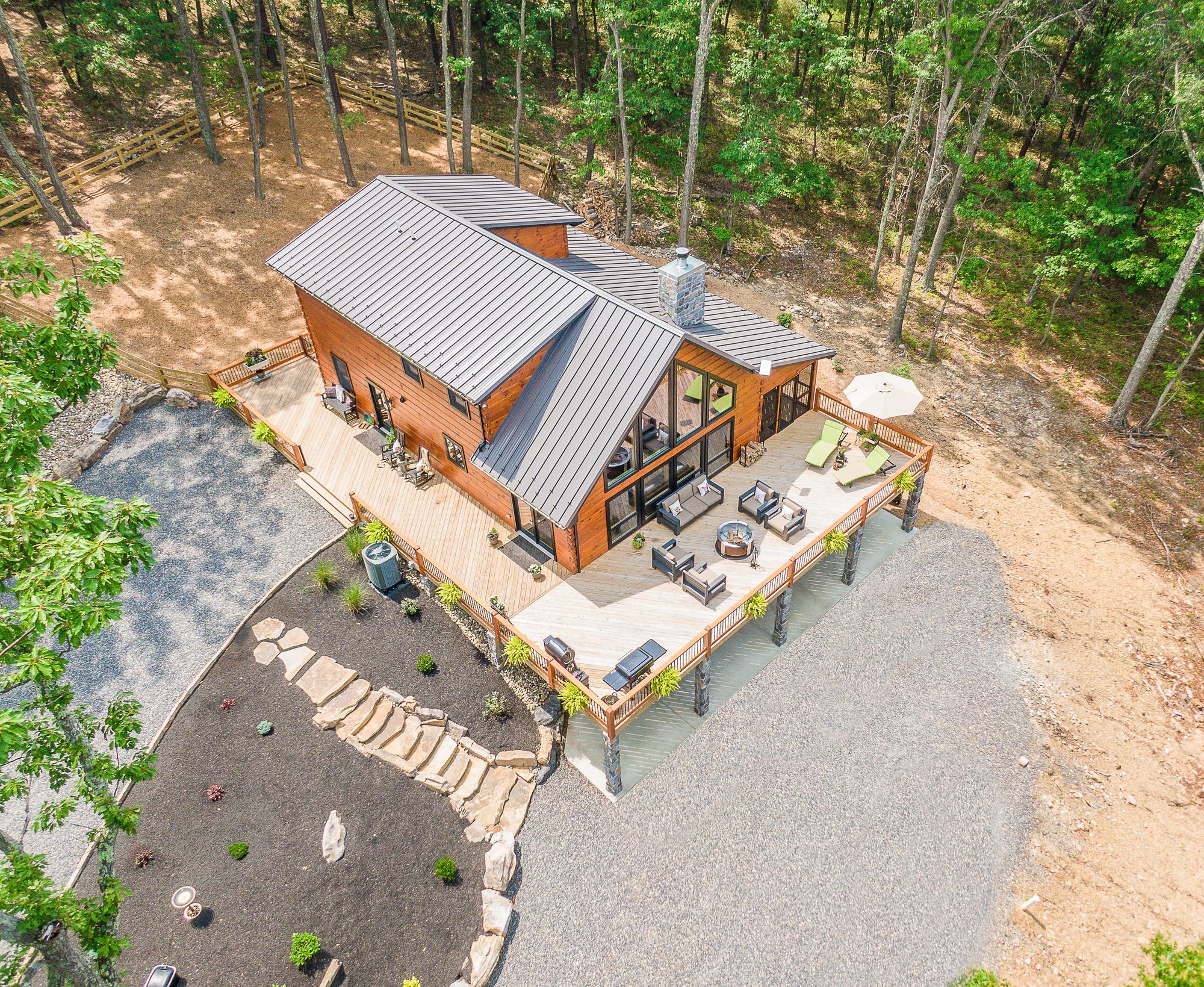 Elevated view looking down on the metal roof and wraparound deck, with cleared ground at the forest edge