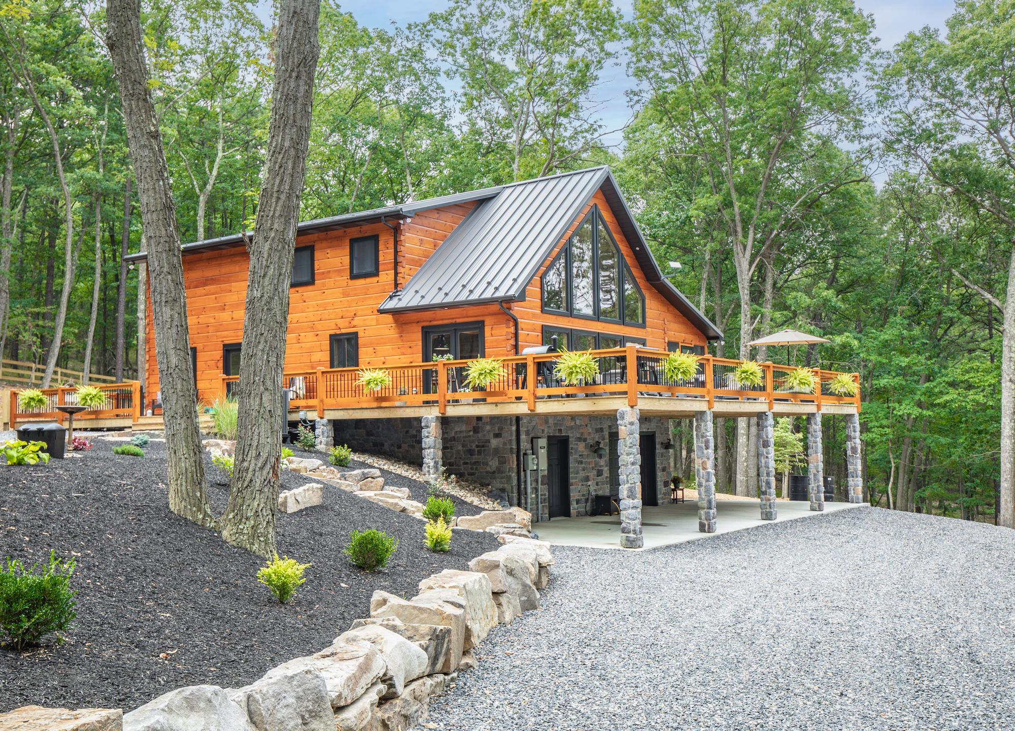 Ground-level view of the log home above a boulder retaining wall and mulched plantings, its deck wrapping the corner