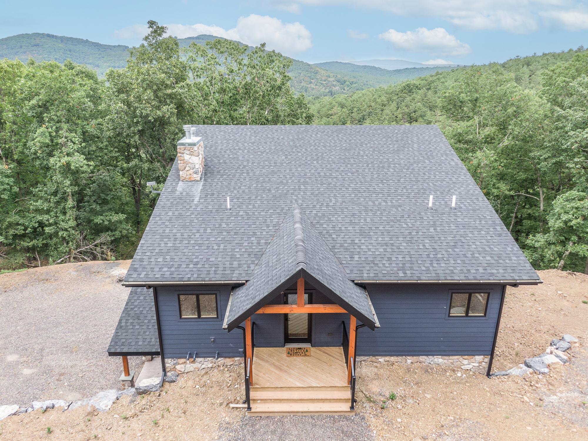 The home from in front and above, its shingle roof and stone chimney set against layered wooded ridges