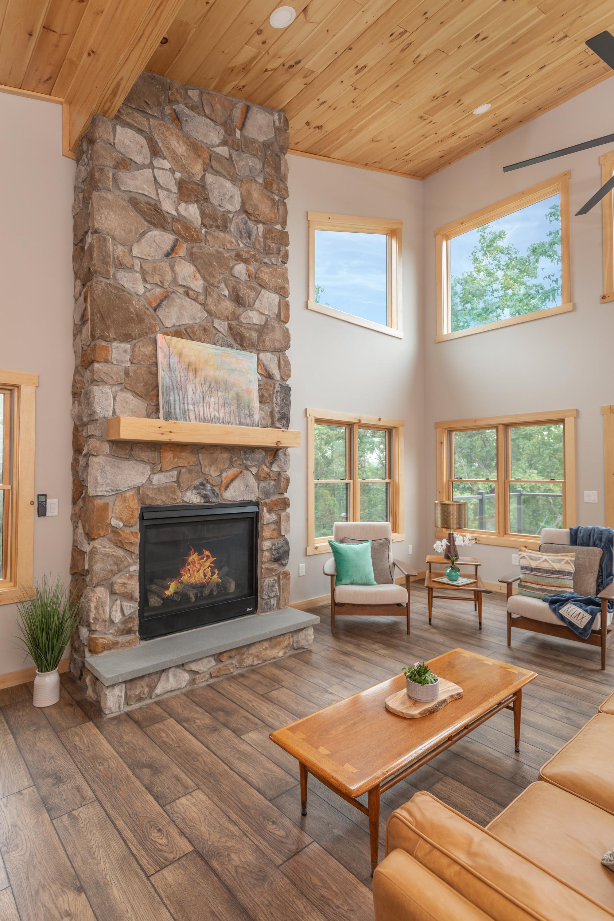 Tall stone fireplace with a lit fire and a timber mantel, beneath a vaulted pine ceiling