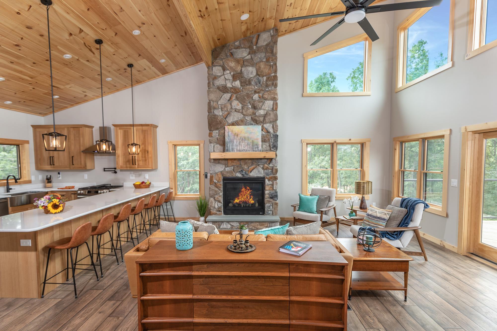 The great room seen across the kitchen island, a lit stone fireplace rising to the vaulted pine ceiling under clerestory windows
