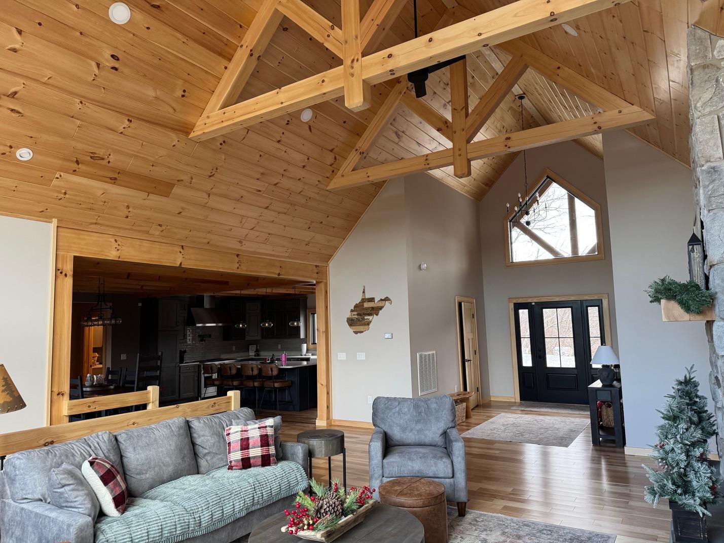 Great room under heavy timber trusses and a pine ceiling, open through to the kitchen, with a stone chimney at the right