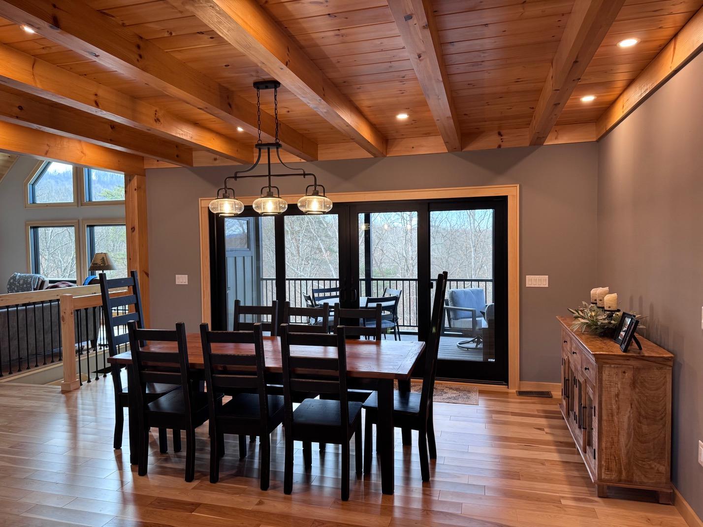 Dining area beneath a beamed ceiling and a linear iron chandelier, with sliding doors onto a screened porch