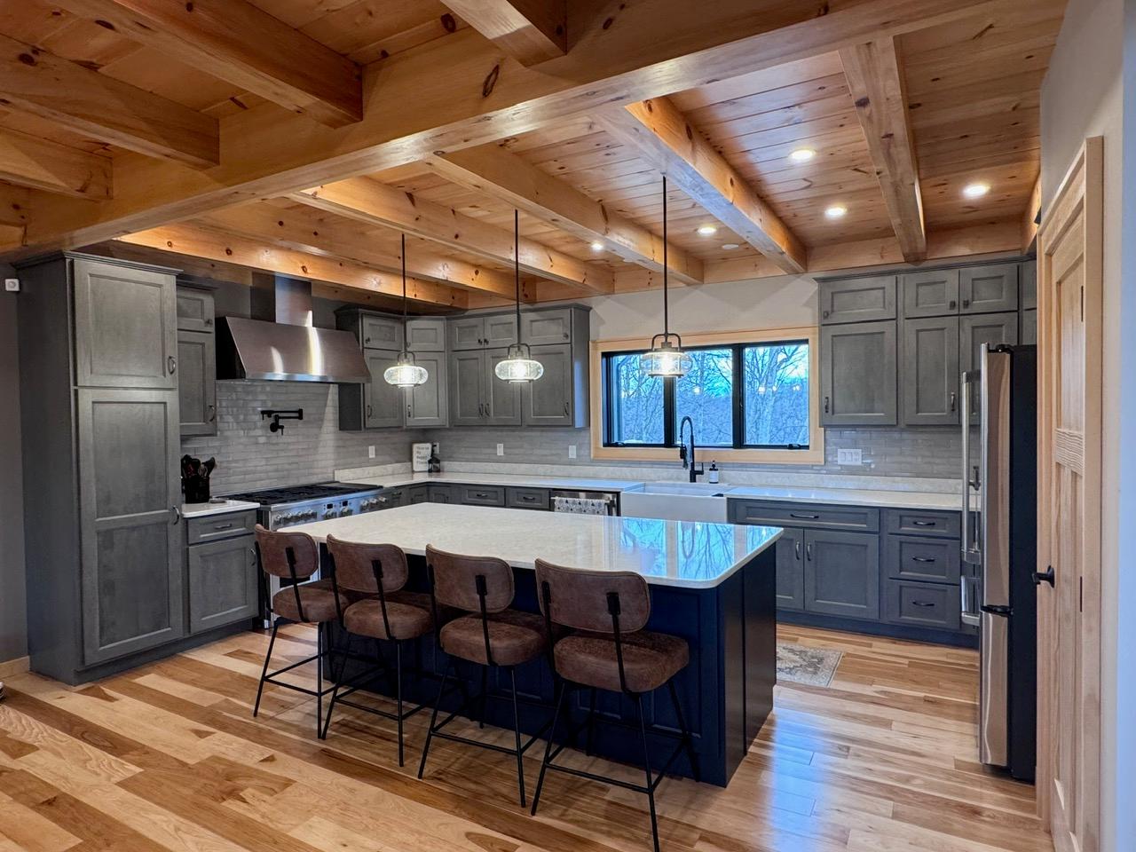Kitchen with gray cabinets, a quartz island and leather stools, beneath an exposed beam ceiling
