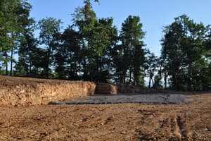 The pad from the driveway side, cut into the slope with a raw bank behind it and summer woods all around.