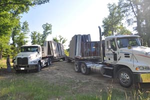 Two flatbed trucks parked on a grassy track, each loaded with upright precast foundation panels, a worker standing beside the nearer one.