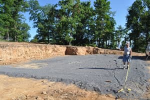 A crew member runs a tape measure across the leveled gravel pad, with the excavated bank and a line of trees behind.