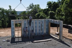 The first basement walls stand on the gravel pad as the crane lowers a panel with a door opening into the corner.