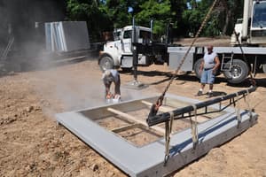 A crew member cuts an opening in a precast wall panel lying on the ground, dust in the air and the crane truck behind.