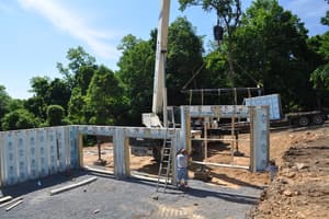 Precast basement walls rising around the gravel pad, the crane boom overhead and a panel with a doorway hanging in place.