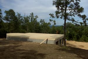 The foundation capped with a plywood subfloor deck, a plank ramp leaning against the wall and a walkout opening below at the right.