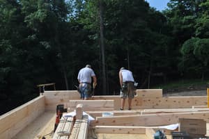 Two of the crew stand among the first courses of square-cut log laid out on the new subfloor, tools and plans at hand.