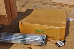 A carton of 12-inch zinc lag bolts open on the subfloor, a shrink-wrapped bundle of them beside it, used to pin the log courses.