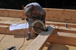 A crew member trims a square-cut log with a circular saw on the subfloor, the building plans spread out beside him.