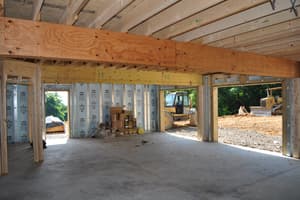 Inside the lower level: a concrete slab, a built-up beam carrying the floor joists overhead, and open doorways looking out to earthmoving equipment.