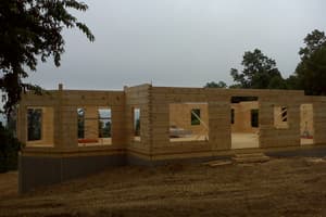 Log walls stacked to full height on the foundation, window and door openings cut through them, under a flat gray overcast sky.
