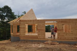 Two men on the deck of a half-built house at log-wall height, one sweeping with a push broom, the other standing in a door opening with the wooded ridge behind him.