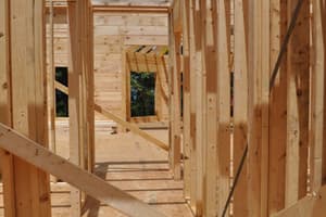 A framed hallway on the main floor, stud partitions running toward log walls beyond and heavy beams crossing overhead against the sky.