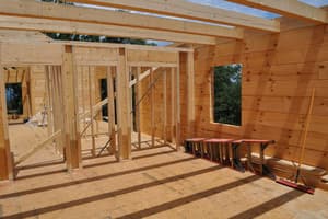 The main floor from inside: a log exterior wall with a window opening cut in it, framed partitions to the left, a folded ladder and a push broom.