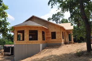 Log walls stacked to the eaves on a gray foundation, window openings still empty, a shingled hip roof over the corner bay and bare dirt all around.