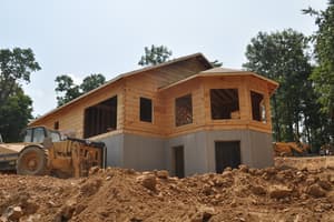 Seen from a heap of excavated clay, log walls and a wide roof overhang rise above a gray foundation with door openings, a loader parked alongside.