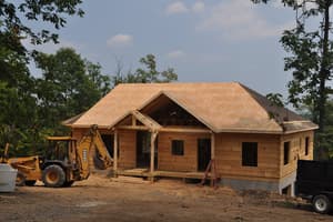 The front of the log home with fresh roof sheathing, a timber-braced porch gable, empty window openings, and a backhoe parked at the near corner.