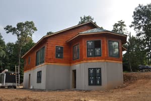 Log walls stained a warm reddish brown with windows now installed and labels still on the glass, above a gray foundation with a walkout door.
