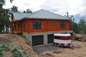 A gable end above two garage bays in the foundation level, standing-seam metal roof finished and windows set, a trailer parked on the gravel.