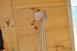 Interior detail of a log wall where a notched pocket lets a bundle of white electrical cables run down beside a bolted upright post.