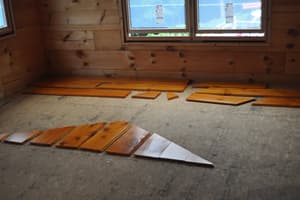 Finish flooring planks laid out loose on the panel subfloor of a log-walled room, sorted for fit below a window that is already set.